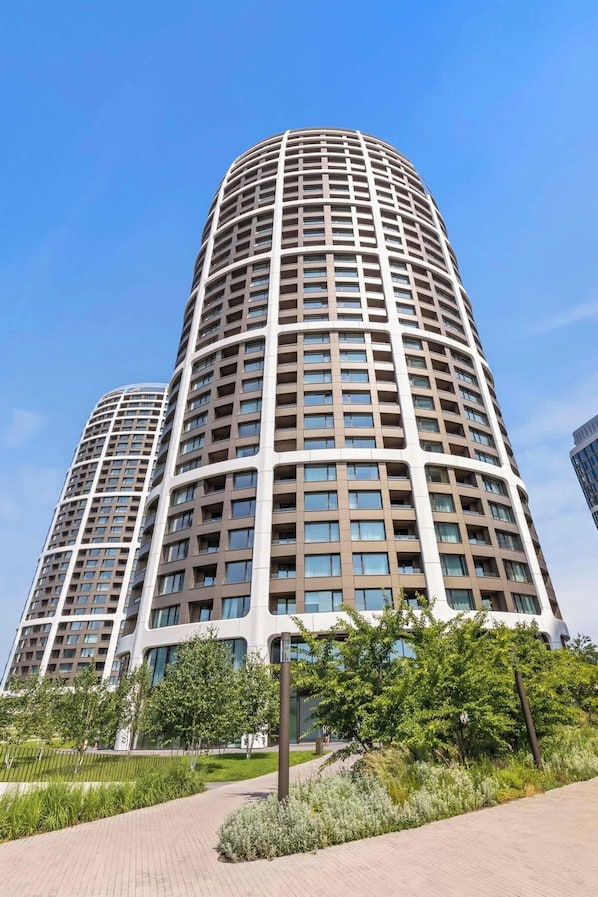Un immeuble moderne de grande hauteur, doté d’une façade incurvée, de nombreux balcons et d’un espace paysager agrémenté d’arbres et d’un sentier.