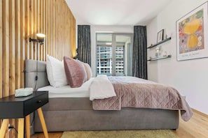 Une chambre à coucher moderne avec un lit, une table de chevet, une lampe et une vue sur un immeuble par la fenêtre.
