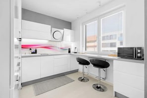 Una cocina moderna con armarios blancos, dos taburetes negros, un microondas y una ventana con persianas.