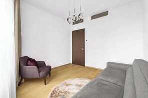 A living room with a grey sofa, a purple armchair, a wooden floor, a door, and a ceiling light fixture.