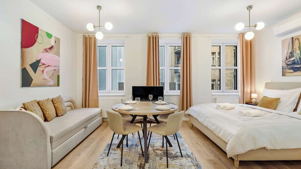 A modern hotel room with a sofa, a bed, a dining table with chairs, a TV, and large windows with curtains. - On the Main Shopping Street - City Center (Bratislava)