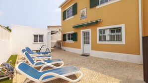 Un edificio con fachada amarilla, contraventanas verdes y un patio con azulejos y sillas de tomar el sol.