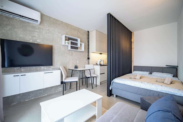 A modern hotel room with a flat-screen TV, a dining area with chairs and a table, a bed with a grey headboard, and a large window with a curtain.