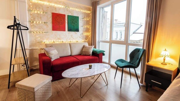 Una sala de estar moderna con un sofá rojo, una mesa redonda de centro, un sillón verde y una pequeña mesita de noche con una lámpara.
