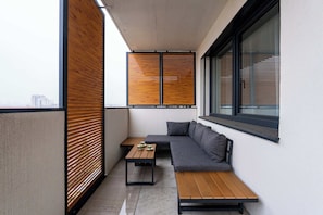Ein Balkon mit einer grauen Couch, Holztischen und einem Fenster mit Jalousien.