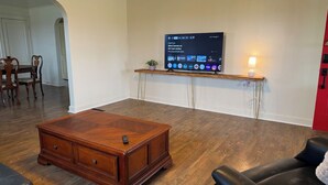 A living room with a wooden coffee table, a black leather sofa, a TV on a stand, and a dining area in the background. - Lucky Horseshoe Farmhouse (Wilmington)