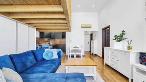 Uma sala de estar moderna com um sofá azul, vigas de madeira no teto, um espaço de jantar branco e uma cozinha ao fundo.