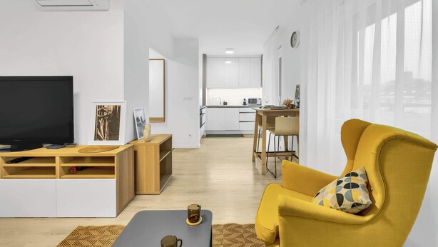 Ein modernes Wohnzimmer mit Fernseher, Unterhaltungseinheit und einem gelben Sessel. Im Hintergrund ist eine Küche zu sehen.