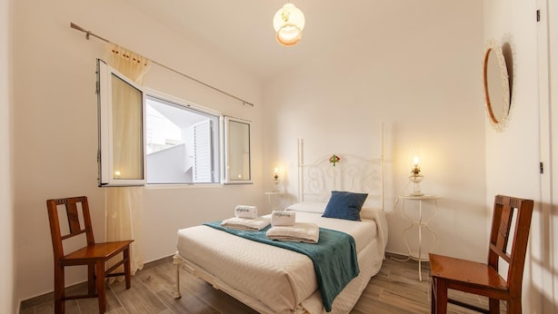 Un dormitorio con una cama, dos sillas, una mesita de noche con lámpara y una ventana con cortinas.