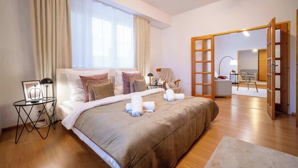 A hotel room with a large bed, a bedside table with a lamp, a chair, a view of the living area, and a wooden door. - Your City Center Stylish & Spacious Apartment (Košice)