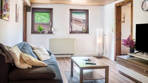 Ein Wohnzimmer mit einer Couch, einem Couchtisch, einem Fernseher und zwei Fenstern mit Pflanzen.