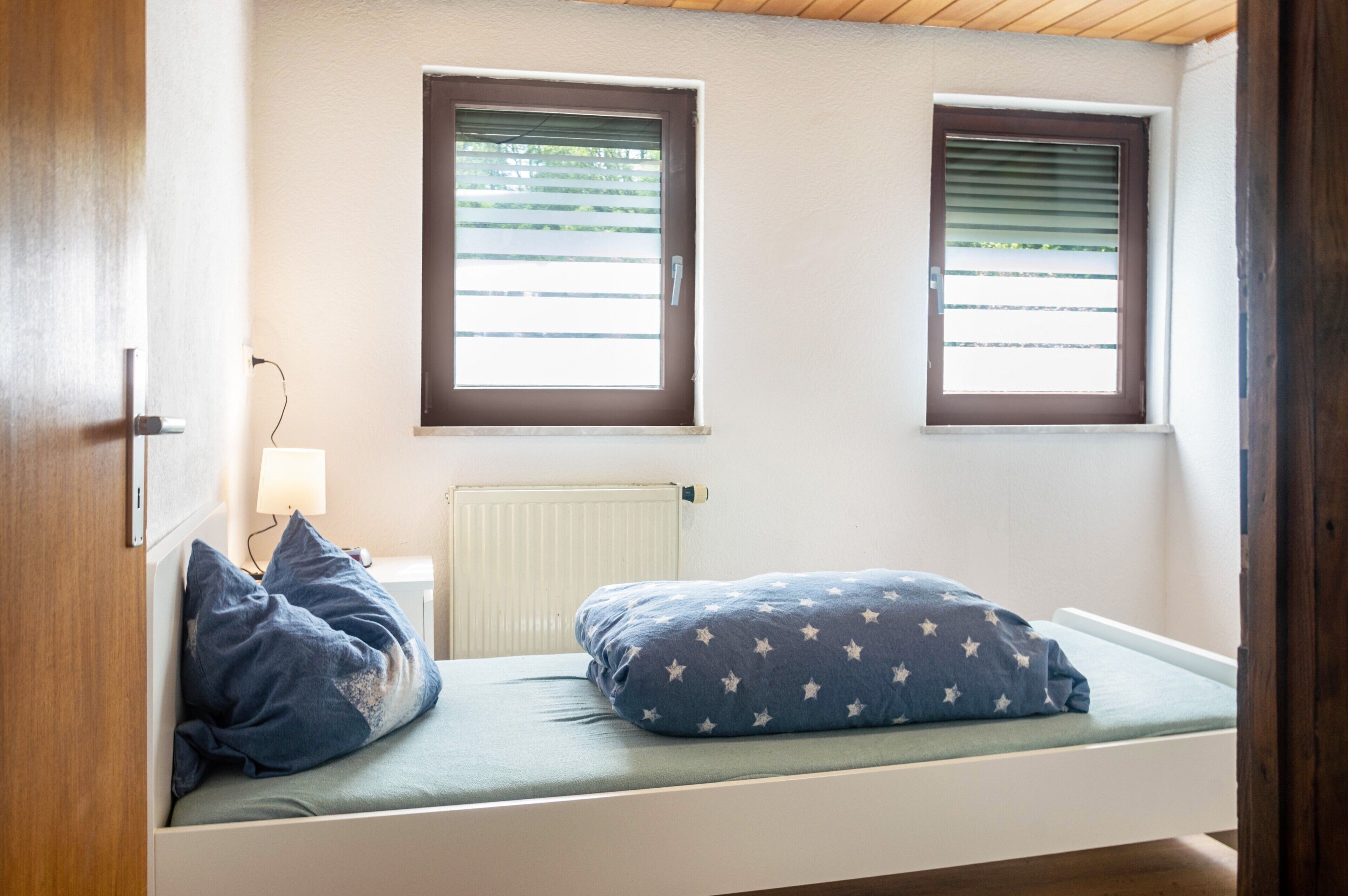 Ein kleines Zimmer mit einem Einzelbett, einem Fenster, einer Heizung und einer Lampe.