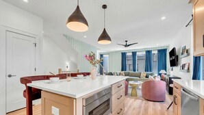 A modern kitchen with a central island, a dining area with a red chair, a sofa, and a ceiling fan.