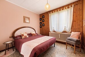 Una camera da letto con testiera in legno, un letto con copripiumino bicolore rosso e beige, un comodino, una sedia, un termosifone e una finestra con tende.