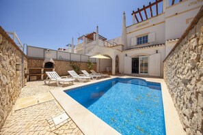 Une piscine avec des chaises longues, un mur en pierre et un bâtiment à la façade blanche ornée d’arcades.