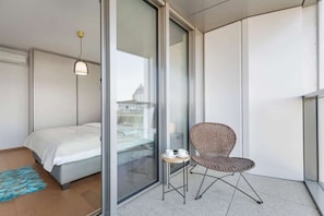 A modern hotel room with a bed, a wicker chair, a small table with cups, and a view through a glass door. - Eurovea Tower Apt 2 Sky View (Bratislava)