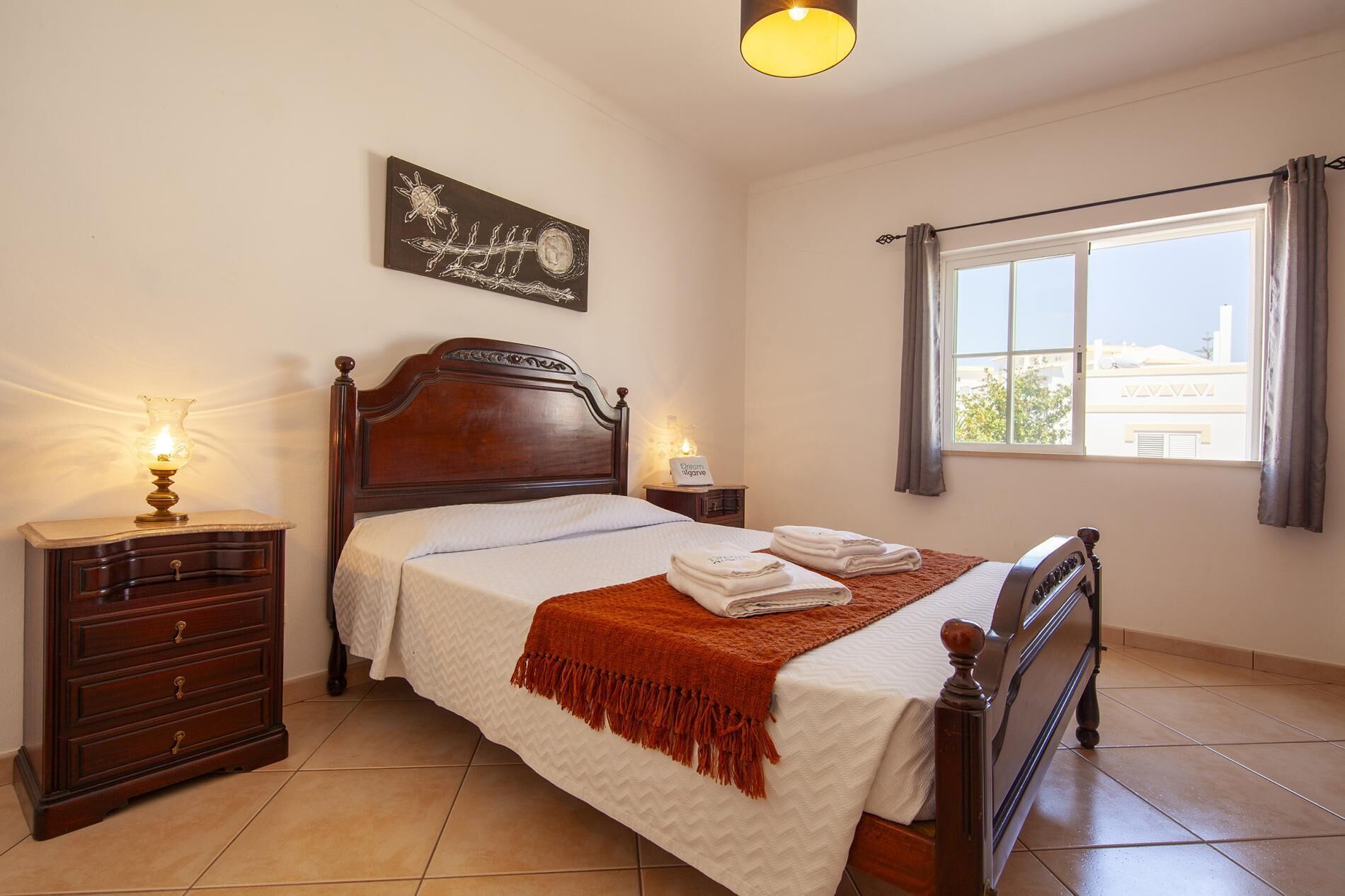 A neatly made bed with a white bedspread and a brown fringe throw, a wooden nightstand with a lamp, a wall-mounted artwork, and a window with curtains.