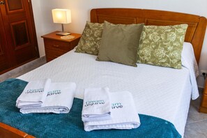 Cama bem arrumada com duas toalhas dobradas, cabeceira de madeira e mesa de cabeceira com abajur.