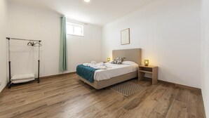 Ein Schlafzimmer mit Bett, Nachttisch, Lampe, Fenster mit Vorhängen und Holzboden.