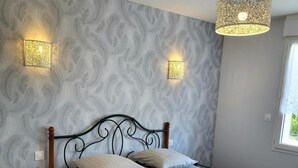 A bed with a metal frame, two wall-mounted lights, patterned wallpaper, and a window with a curtain. - Gîte Cadet Rousselle: private terrace, private garden & Wi-Fi in Lessay (Lessay)