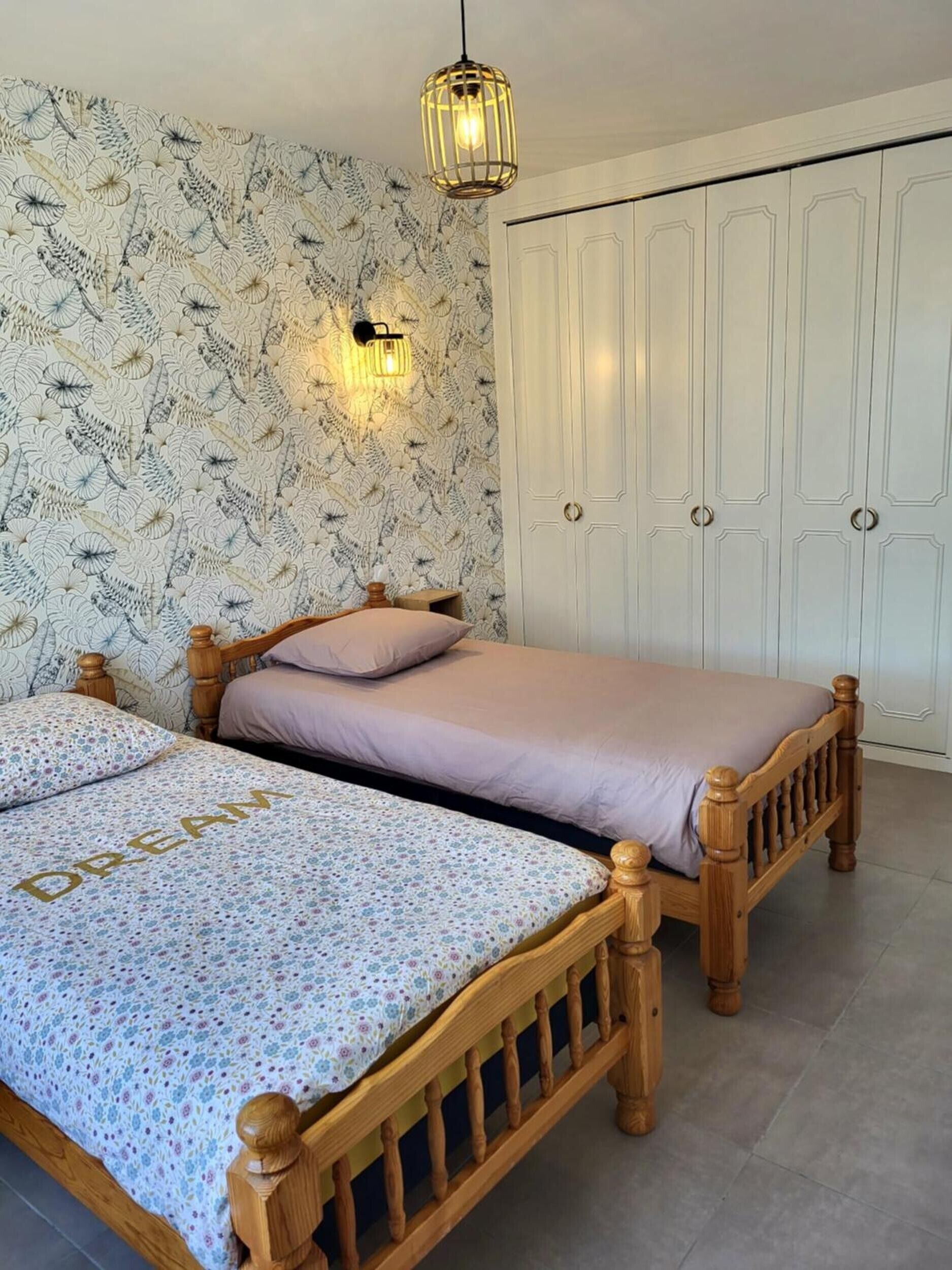 Un dormitorio con dos camas de madera, un papel tapiz estampado, un armario blanco y una lámpara colgante.