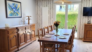 A dining area with a wooden table set for four, flanked by wooden cabinets, a television on the wall, and a large window with a view of greenery. - Gîte Cadet Rousselle: private terrace, private garden & Wi-Fi in Lessay (Lessay)