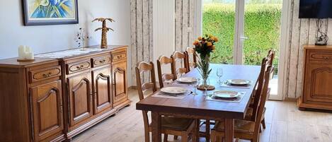 Un coin repas avec une table en bois dressée pour quatre personnes, bordée de meubles en bois, un téléviseur fixé au mur et une grande fenêtre donnant sur un paysage verdoyant.
