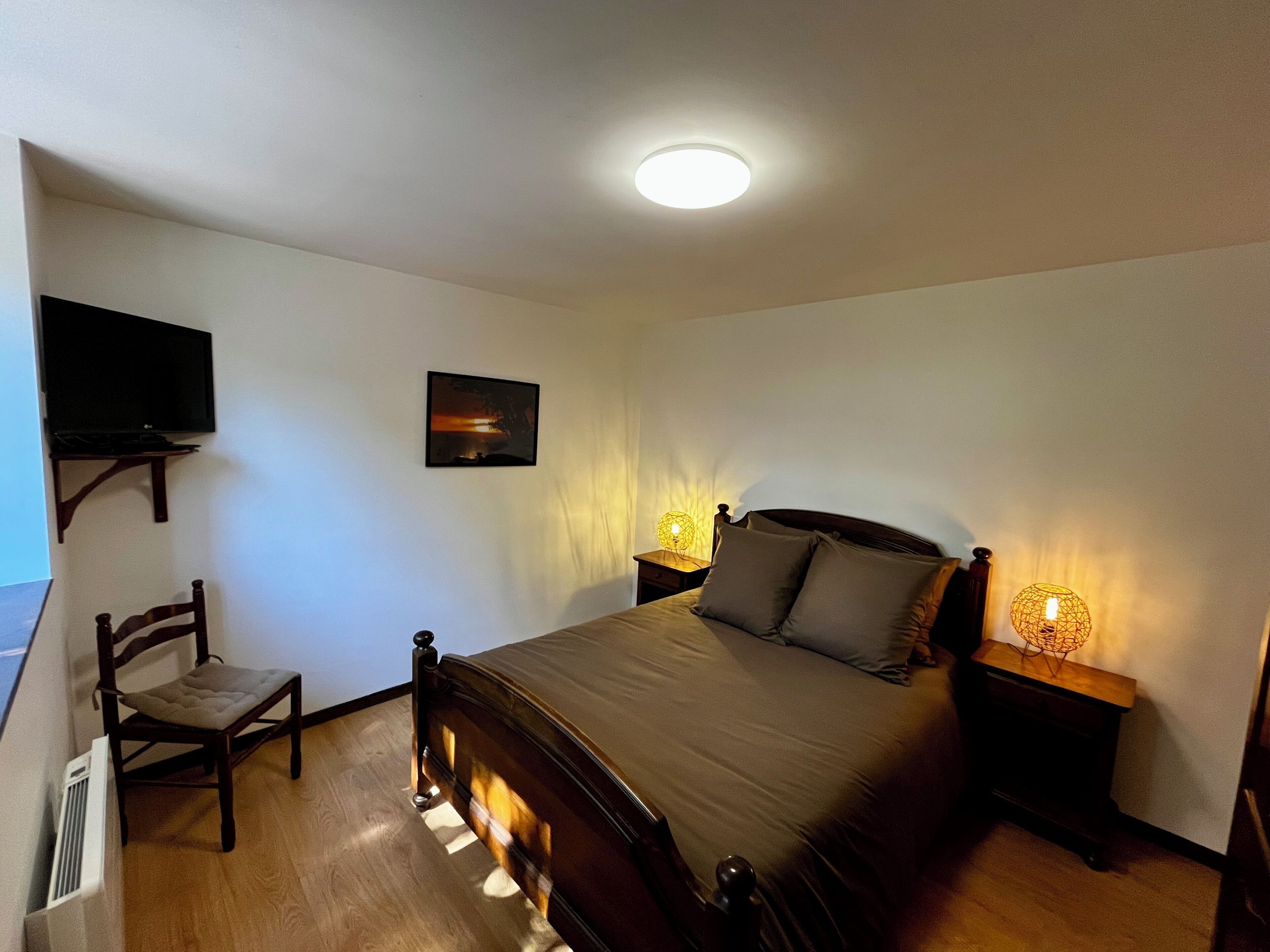 Een slaapkamer met een bed, een stoel, een aan de muur bevestigde televisie en een schilderij.