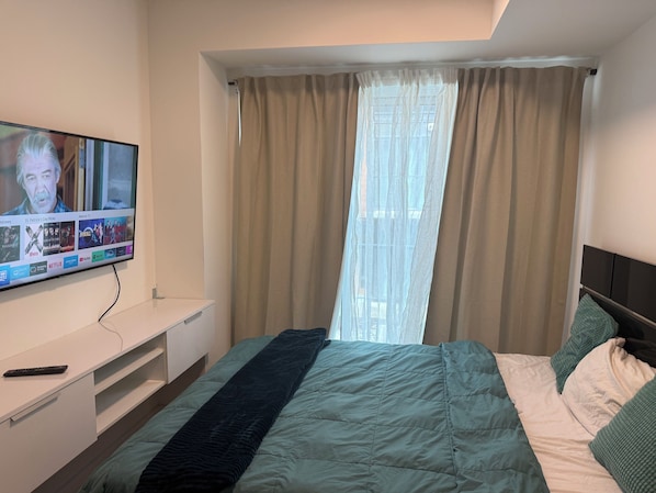 Ein Schlafzimmer mit einem Bett, einem an der Wand montierten Fernseher, einem Fenster mit Vorhängen und einer Fernbedienung auf dem Nachttisch.
