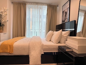 Ein Schlafzimmer mit einem Bett, einem Nachttisch mit Büchern, einem Fenster mit Vorhängen und einem an der Wand befestigten Fernseher.