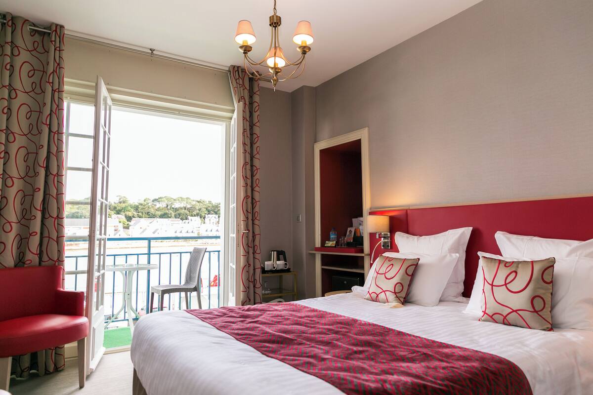 A hotel room with a bed, a red chair, a small table, a chair, a nightstand, a lamp, and a view of the outside.