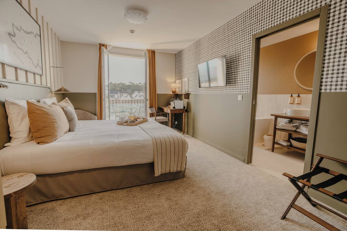 A bedroom with a bed, a desk, a chair, a TV, and a bathroom with a mirror and sink.