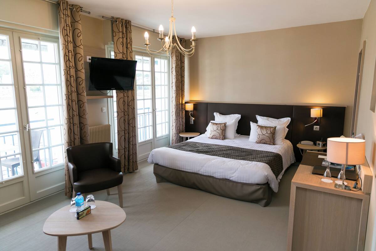 A hotel room with a large bed, a TV mounted on the wall, a chair, a small table, and a bedside lamp.