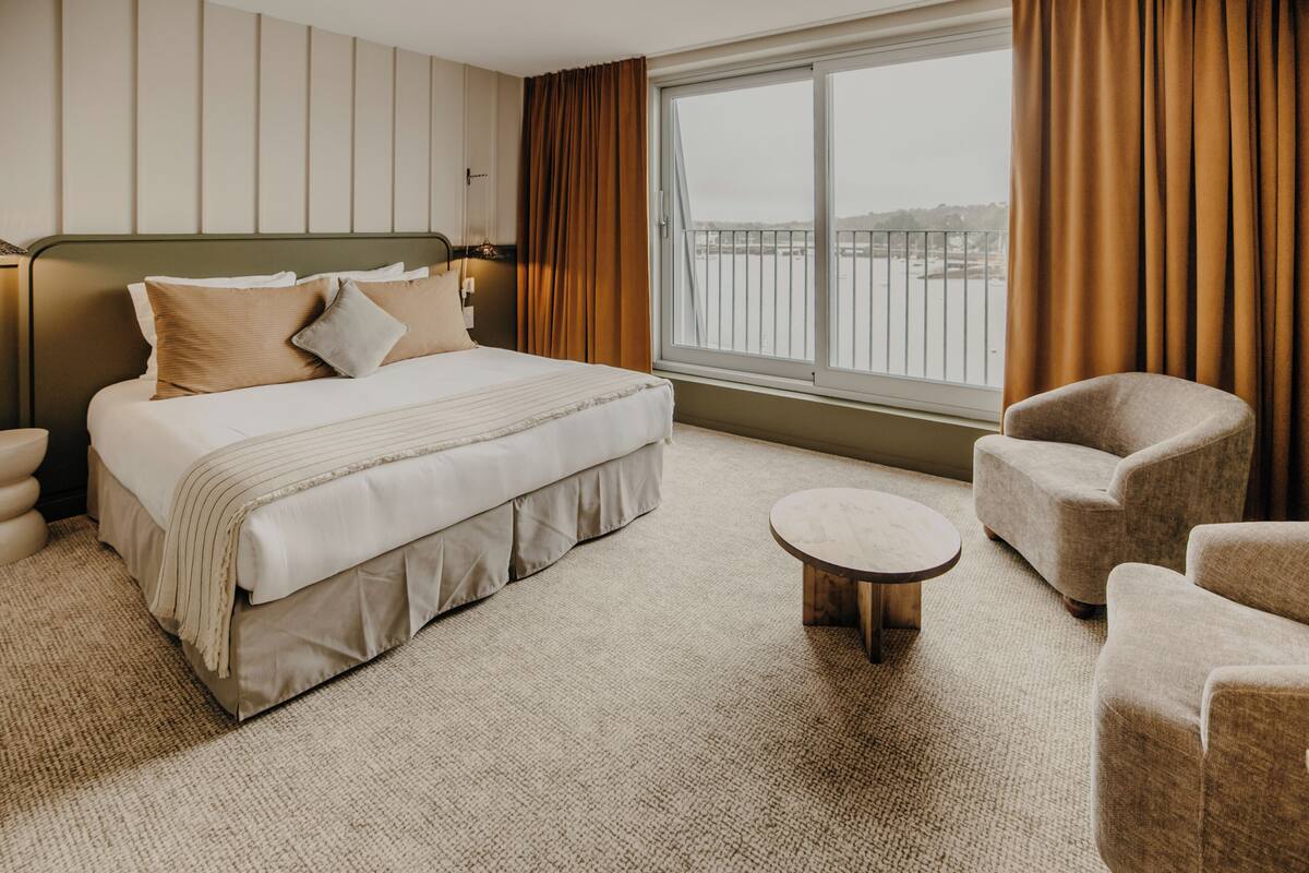 A hotel room with a bed, a round table, two armchairs, a window with curtains, and a view of a body of water.