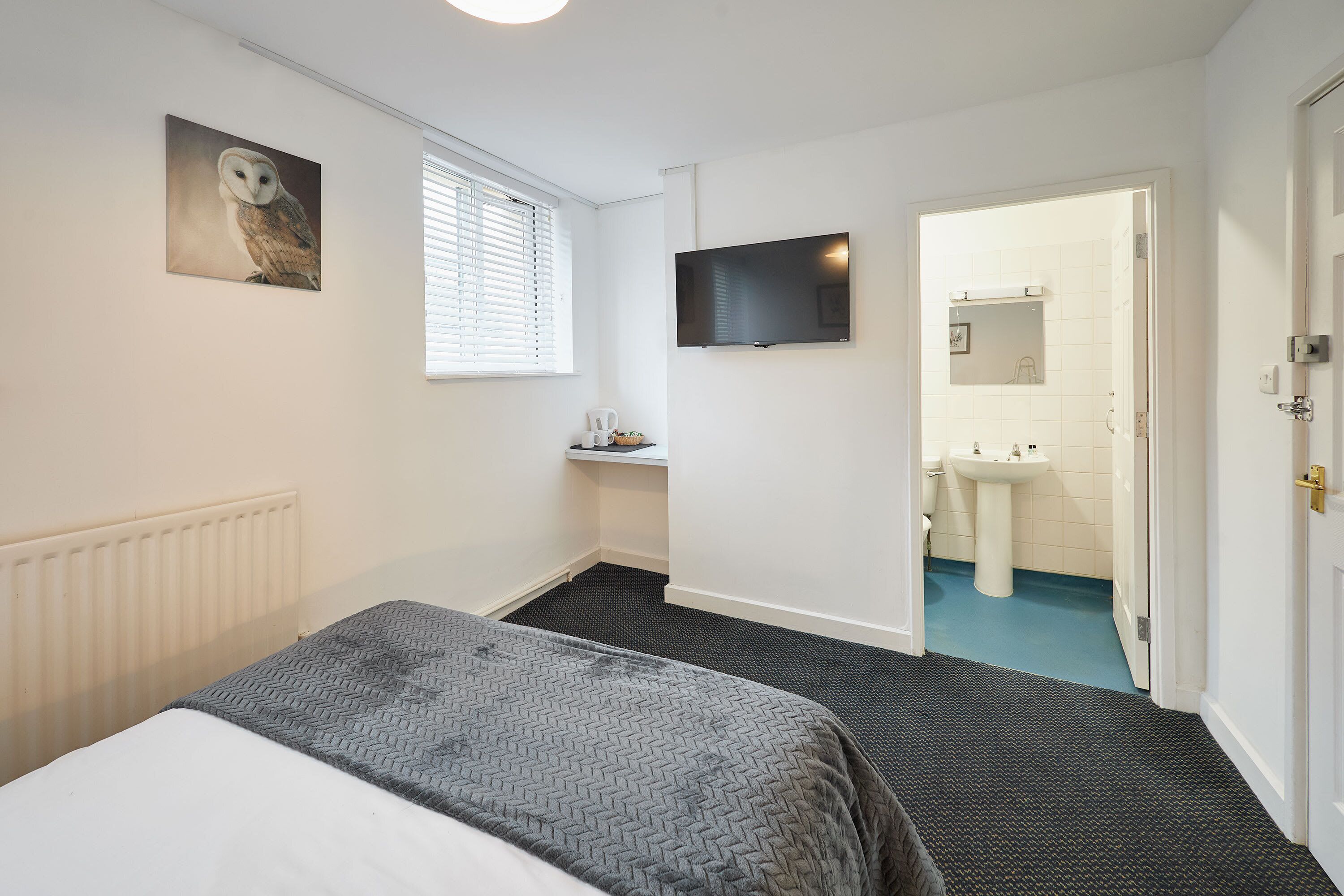 Ein Schlafzimmer mit Bett, Fernseher, Gemälde, Heizkörper, Fenster und einem durchgehenden Türbogen sichtbarem Badezimmer.