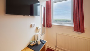 Hotelkamer met flatscreen-tv, raam met rode gordijnen, radiator en een klein tafeltje met koffiezetapparaat en een kom met snacks.