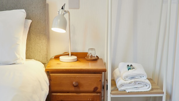 Een net opgemaakt bed met witte lakens, een bedlampje, een houten nachtkastje met een lade, en een plank met opgevouwen handdoeken.