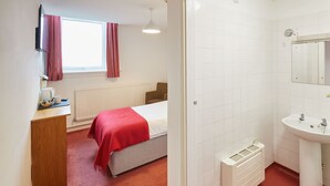 Een hotelkamer met een bed, een bureau met waterkoker, een radiator, een witte wastafel en een raam met gordijnen.