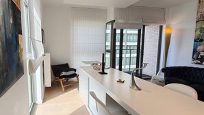 Una stanza moderna con una scrivania bianca, un divano nero, un quadro astratto di grandi dimensioni e vista su un edificio attraverso la finestra.