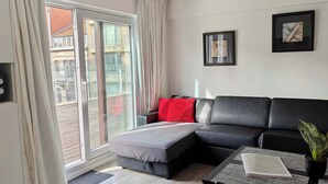 Ein modernes Wohnzimmer mit einer schwarzen Ledersofa, einem roten Sitzkissen, einem kleinen Tisch mit einem Buch und einer Topfpflanze sowie einer Glastür zum Außenbalkon.