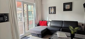 A modern living room with a black leather sofa, a red throw pillow, a small table with a book and a potted plant, and a sliding glass door leading to an outdoor balcony. - Beachfront Knokke Apt w/ Panoramic Terrace Views (Knokke-Heist)
