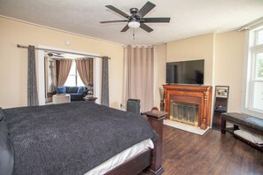 A bedroom with a large bed, a ceiling fan, a fireplace, and a television on the wall.