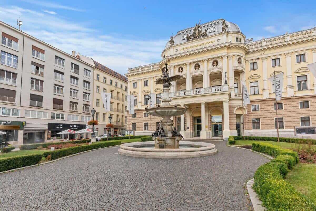 A historic building with a central fountain, surrounded by a cobblestone plaza and modern buildings.