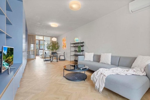 Ein modernes Wohnzimmer mit einem grauen Sofa, einem Glastisch und einem Fernseher links.