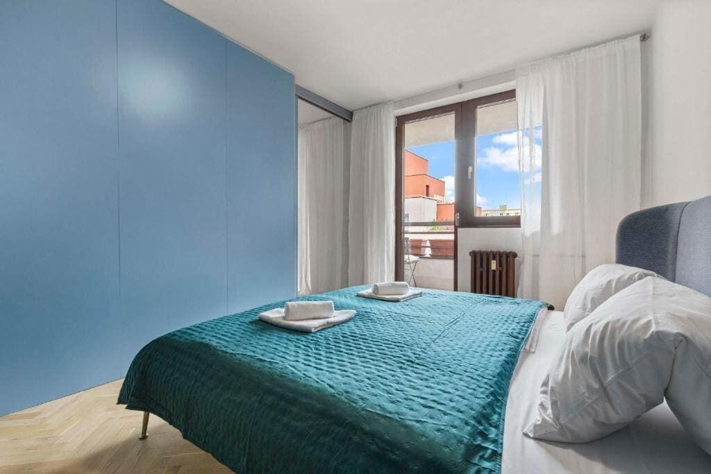 Une chambre à coucher avec un lit, une fenêtre avec des rideaux, une vue sur les bâtiments à l’extérieur et un mur bleu.