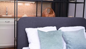 A bed with white and light blue pillows, a dark headboard, and a kitchen area with a sink and cabinets in the background.