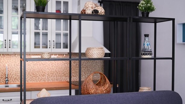 A modern room with a bed, a black metal shelf with baskets, and a kitchen area in the background.