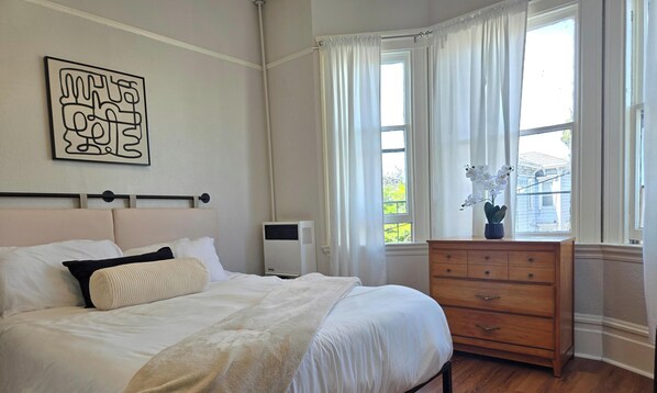 Quarto com cama, cómoda, radiador e janela com cortinas.