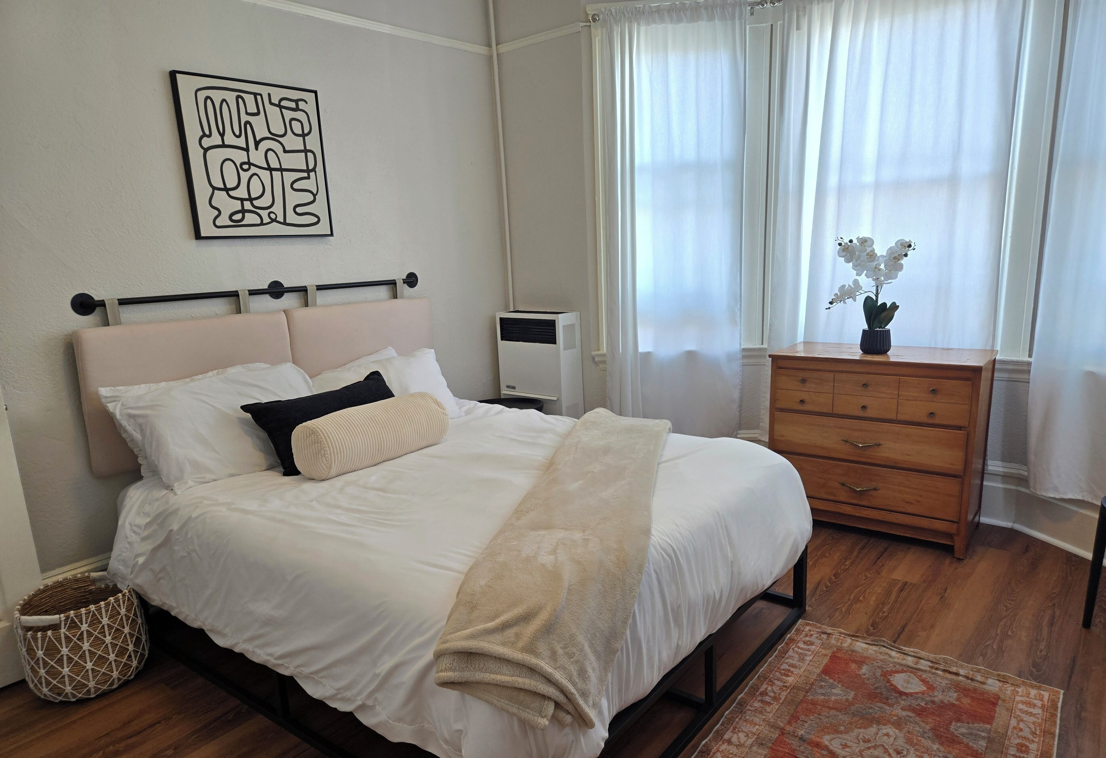 Una cama bien hecha con cabecera, una mesita de noche, un aparador y una obra de arte colgada en la pared.