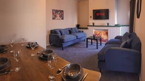 A living room with a fireplace, a sofa, a TV, and a dining table set with wine glasses and plates. - 4 Bed • Parking • Near M4 • Ideal for Workers (Aberavon)
